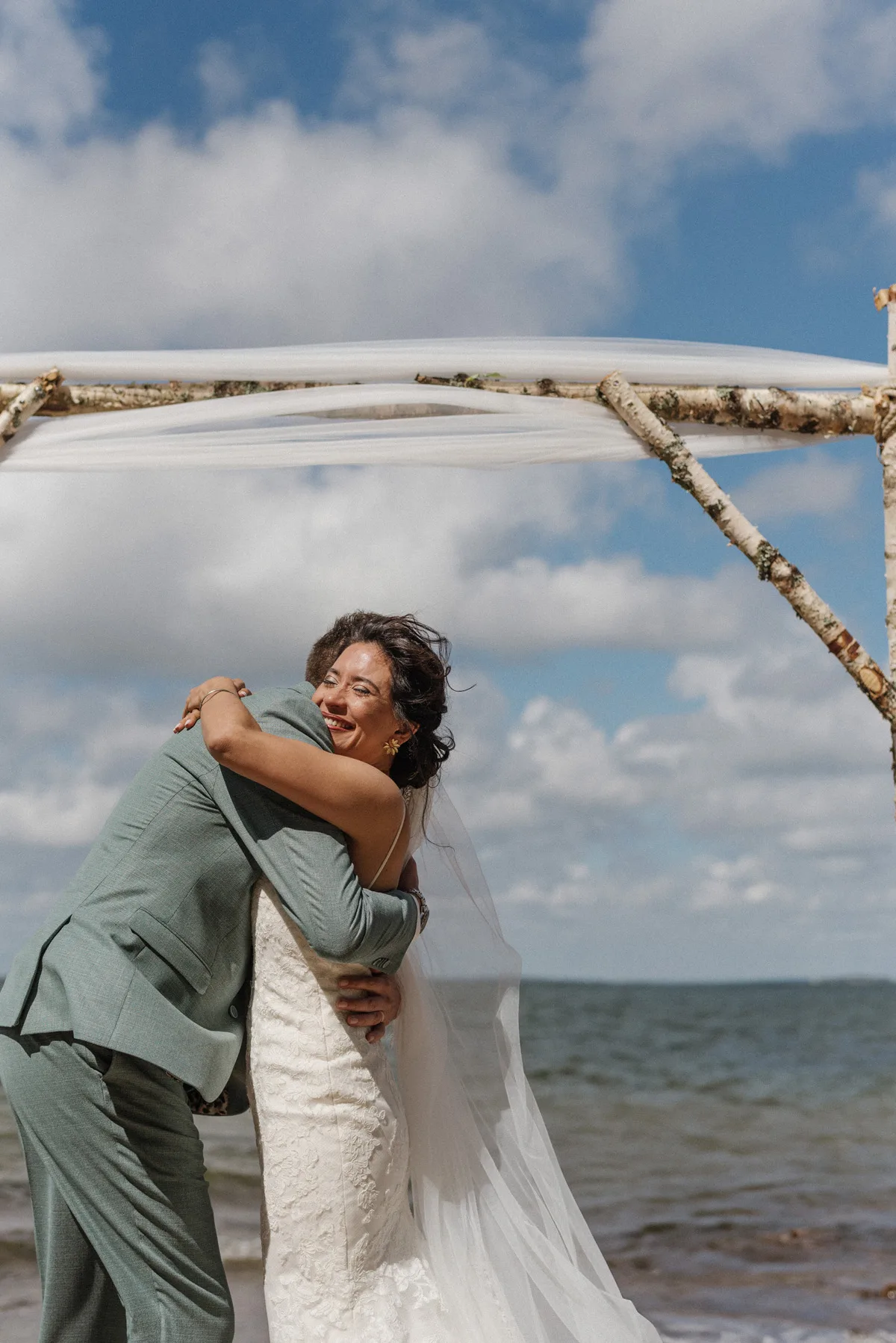 A joyful couple embracing during their seaside wedding ceremony in Finland, with a scenic waterfront background and fluffy clouds in the sky.