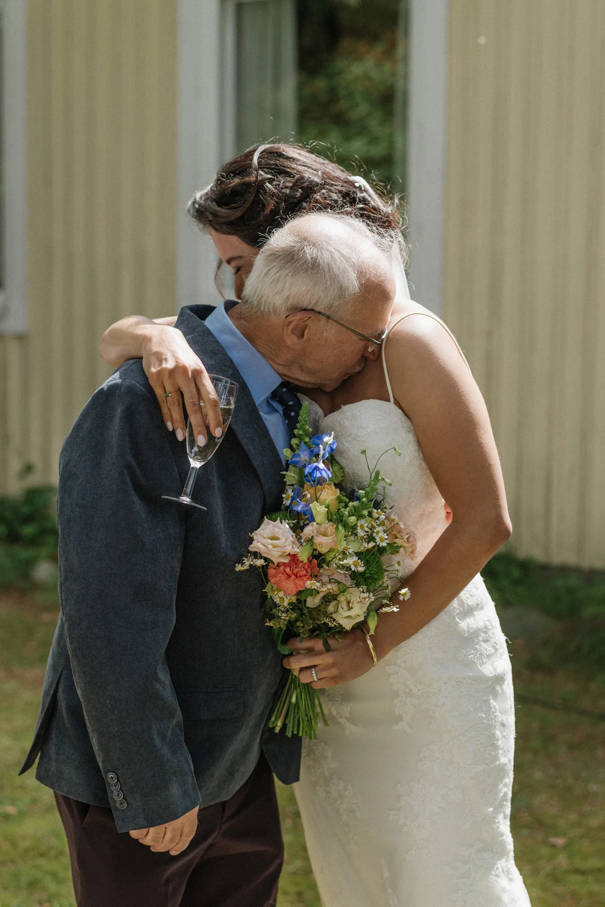 A bride embraces her father in a tender moment during an outdoor wedding ceremony, holding a bouquet of colorful flowers while he kisses her on the forehead.